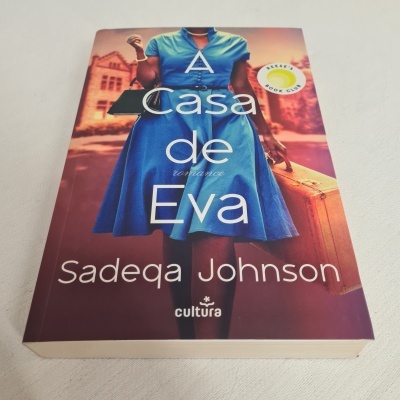 Capa do livro A Casa de Eva com mulher de vestido azul segurando mala castanha
