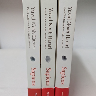 Três livros da série Sapiens de Yuval Noah Harari com capas brancas e base vermelha, numerados e com etiquetas pretas.