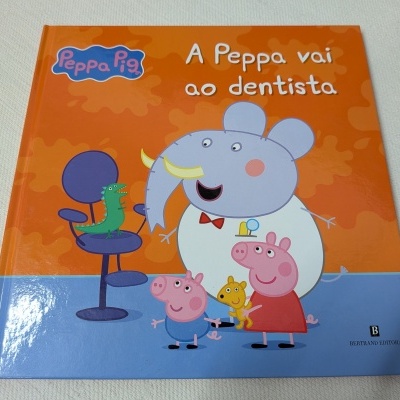 Livro infantil Peppa Pig com título A Peppa vai ao dentista e personagens em fundo laranja e azul.