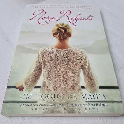 Capa do livro 'Um Toque de Magia' de Nora Roberts com mulher de costas vestindo blusa de renda creme e montanhas ao fundo