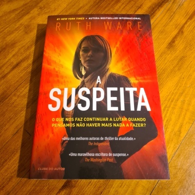 Capa do livro 'A Suspeita' de Ruth Ware sobre superfície de madeira