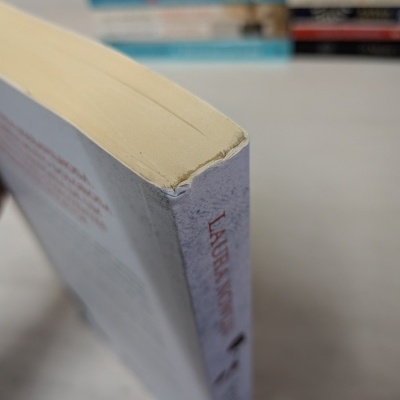 Livro com lombada branca e letras vermelhas, canto superior gasto, vários livros ao fundo