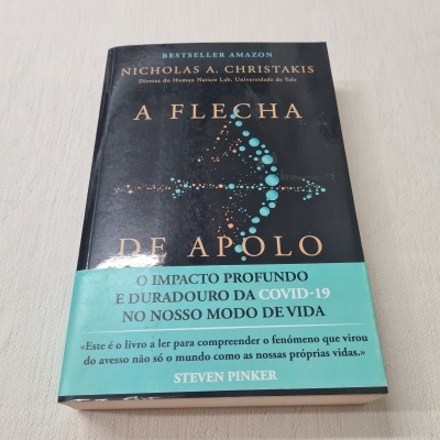 Livro A Flecha de Apolo com capa preta e faixa azul, sobre mesa branca