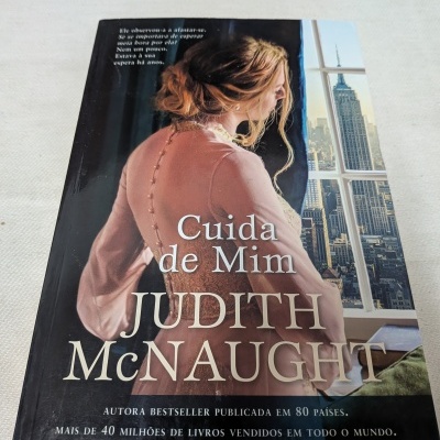 Capa do livro Cuida de Mim de Judith McNaught com mulher de vestido rosa junto a janela com vista de cidade.