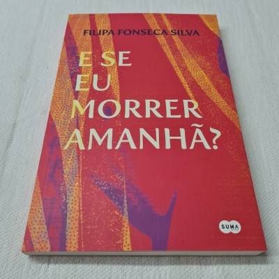 Livro vermelho com texto em amarelo e preto, com padrões laranja e roxo, sobre tecido branco