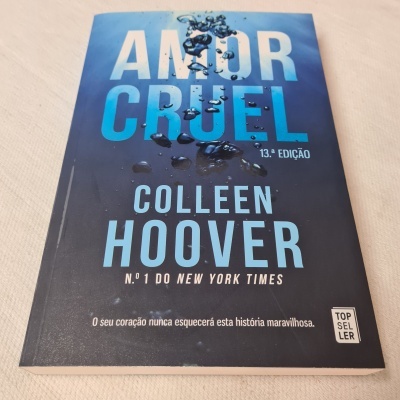 Livro 'Amor Cruel' de Colleen Hoover com capa azul e preta em degradê sobre superfície branca