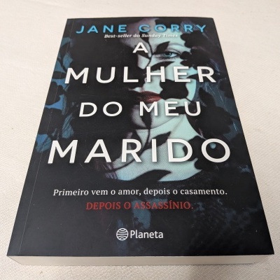Capa do livro 'A Mulher do Meu Marido' com rosto feminino estilizado e texto em branco e vermelho sobre fundo preto