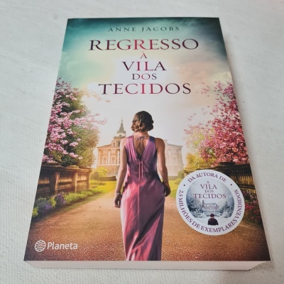 Capa do livro 'Regresso à Vila dos Tecidos' com mulher de costas numa estrada florida
