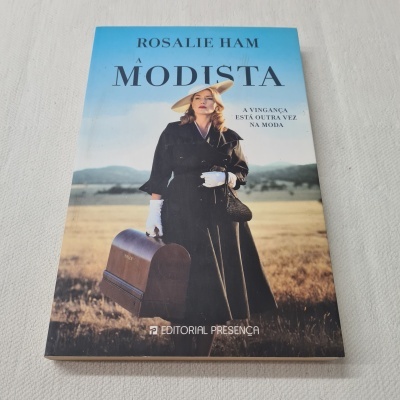 Capa do livro A Modista com mulher vestida de preto e mala castanha em campo aberto.