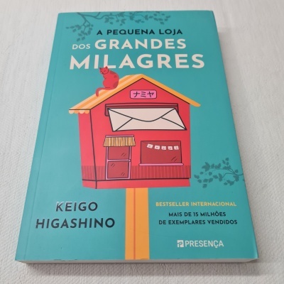 Livro 'A Pequena Loja dos Grandes Milagres' com capa azul e ilustração de casinha vermelha.