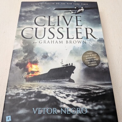 Capa de livro com navio a arder no mar, céu nublado, avião e texto CLIVE CUSSLER por GRAHAM BROWN VETOR NEGRO.