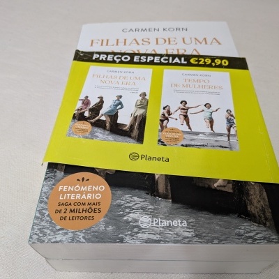 Caixa caixa com dois livros de capa dura, faixa amarela com preço especial €29,90, selo de fenómeno literário, fundo madeira e água.