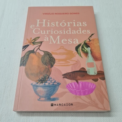 Capa de livro cor de rosa com ilustrações de frutas, peixe e taça, texto 'Histórias e Curiosidades à Mesa'.