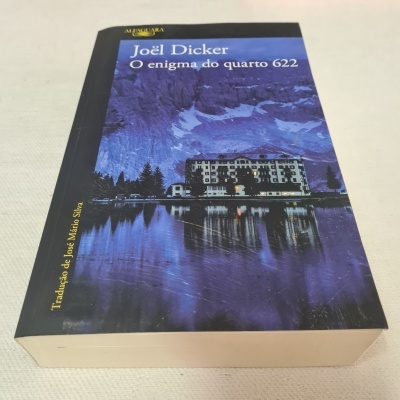 Livro O enigma do quarto 622 de Joël Dicker com capa azul e imagem de edifício e lago