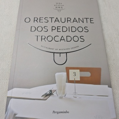 Capa de livro com título O RESTAURANTE DOS PEDIDOS TROCADOS e uma mesa de restaurante simples com itens sobre a toalha branca.