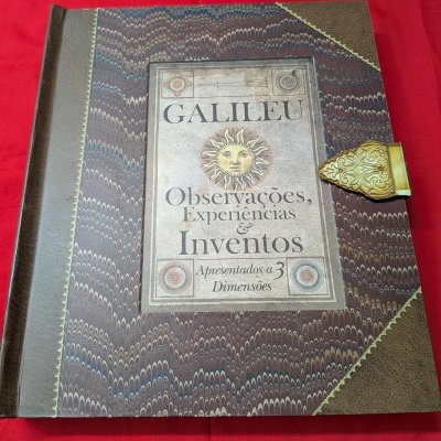 Livro antigo decorativo com fecho dourado e texto na capa sobre tecido vermelho