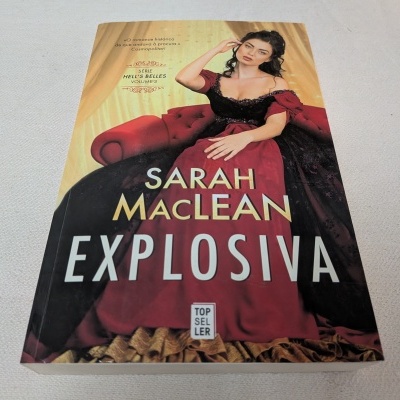 Capa do livro Explosiva de Sarah MacLean com uma mulher em vestido vermelho escuro e preto num sofá vermelho