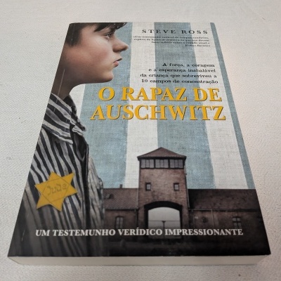 Capa do livro O Rapaz de Auschwitz com jovem em uniforme listrado e portão de campo de concentração ao fundo
