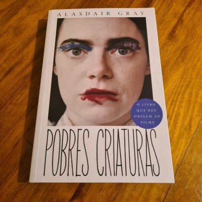 Capa do livro 'Pobres Criaturas' de Alasdair Gray com rosto de mulher e selo azul sobre mesa de madeira