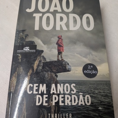 Capa de livro com pessoa de casaco vermelho em rocha, texto João Tordo, Cem Anos de Perdão, thriller.