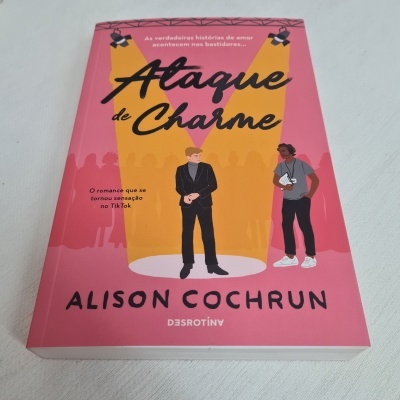Livro 'Ataque de Charme' de Alison Cochrun com capa rosa e amarelo, ilustrações de pessoas