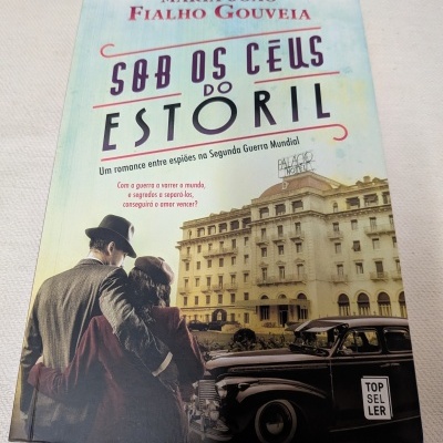 Capa do livro Sob os Céus do Estoril com casal vintage e carro antigo em frente a hotel.