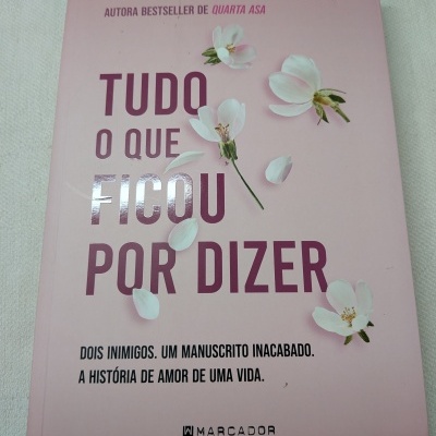 Livro Tudo o que ficou por dizer com capa rosa e flores brancas sobre tecido branco.