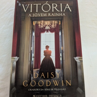Capa de livro intitulada VITÓRIA A JOVEM RAINHA com mulher de vestido preto de costas num corredor.