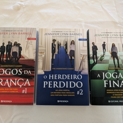 Três livros da série The Inheritance Games em português com capas em vermelho, azul e verde e silhuetas humanas com interrogações.