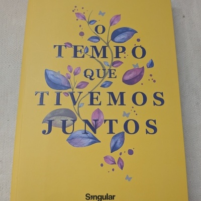 Capa amarela de livro com título O TEMPO QUE TIVEMOS JUNTOS e folhas roxas e azuis