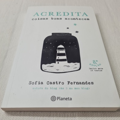 Livro Acredita com capa branca, ilustração de farol num frasco e texto em verde e preto.