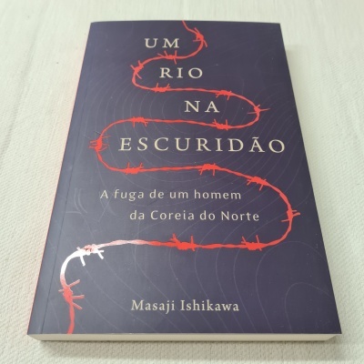 Livro 'UM RIO NA ESCURIDÃO' com arame farpado vermelho na capa azul escura