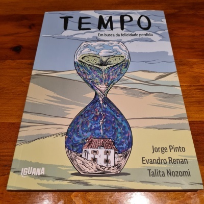 Capa de livro com ampulheta ilustrada, texto TEMPO Em busca da felicidade perdida, autores e selo IGUANA sobre mesa de madeira