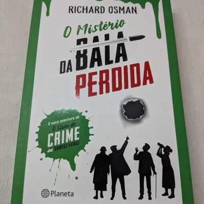 Livro O Mistério da Bala Perdida de Richard Osman com capa verde e branca e silhuetas pretas.