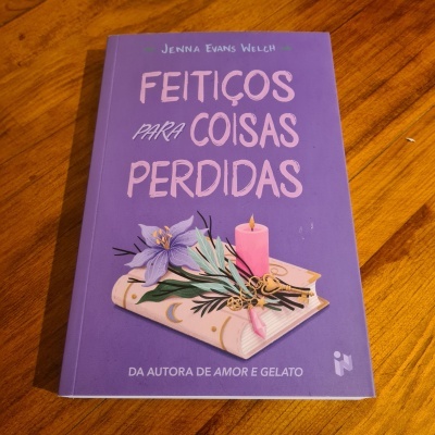 Livro 'Feitiços para Coisas Perdidas' com capa roxa e ilustrações de livro, flor, vela e chaves sobre mesa de madeira.