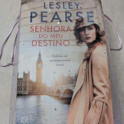 Saco lilás com bolso contendo livro Senhora do Meu Destino com capa que mostra Big Ben e mulher casaco bege.