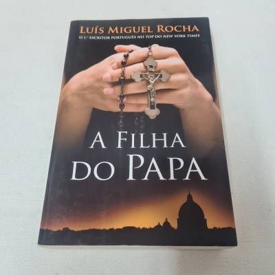 Capa do livro A Filha do Papa de Luís Miguel Rocha com mãos segurando um terço e fundo de silhueta urbana ao pôr do sol