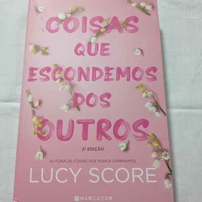 Livro rosa 'Coisas Que Escondemos Dos Outros' com flores brancas sobre tecido branco.