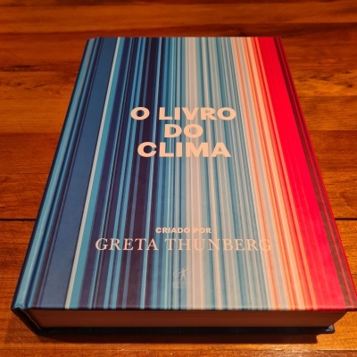 Livro 'O LIVRO DO CLIMA' capa dura com listras azuis, brancas e vermelhas sobre mesa de madeira