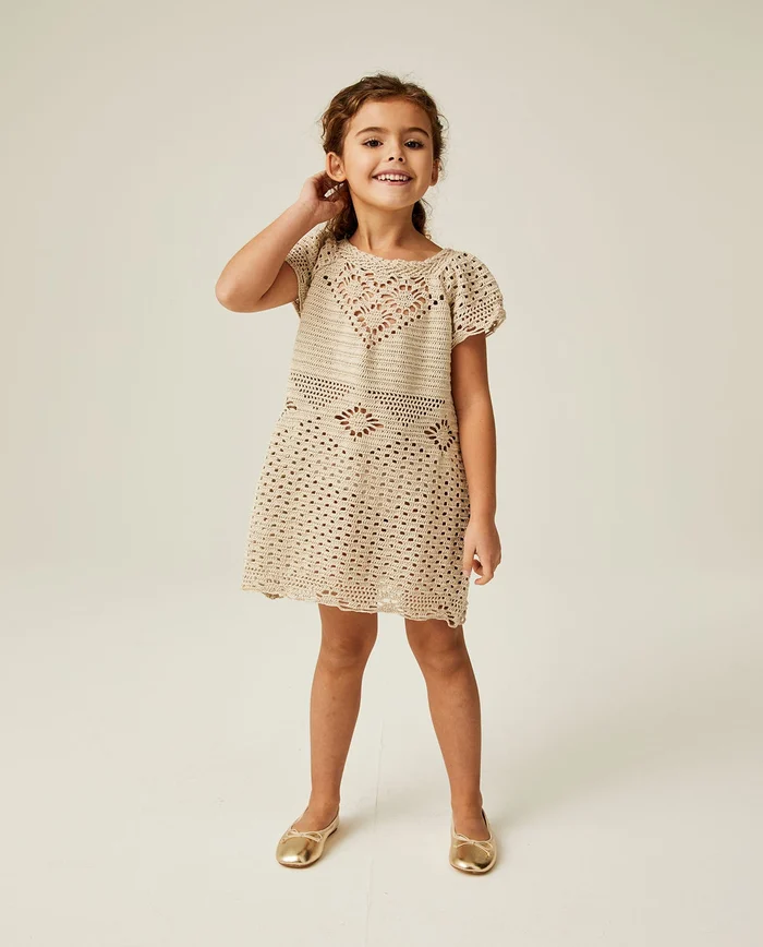 Menina com vestido bege de crochê e sapatos dourados em fundo claro