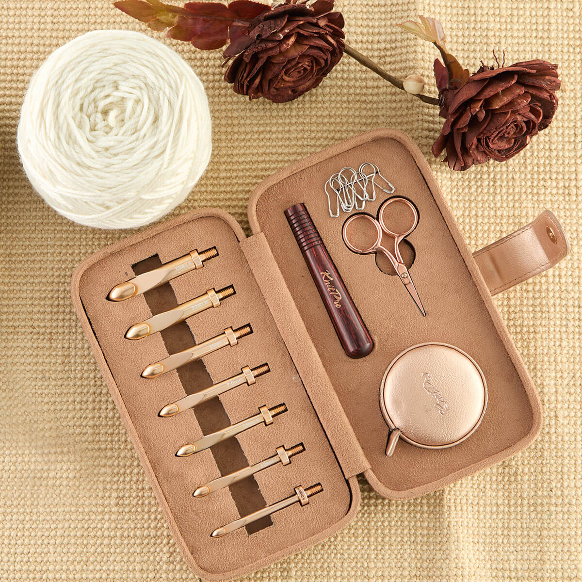 Conjunto de acessórios para trabalho manual com agulhas douradas, tesoura pequena, apontador e case, numa caixa bege sobre tecido texturizado com flores secas