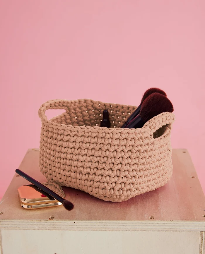 Cesto bege tricotado com pincéis de maquilhagem dentro sobre superfície de madeira e fundo rosa