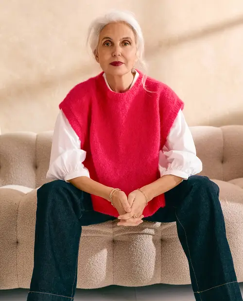 Mulher de cabelo branco com camisola vermelha de malha, camisa branca e jeans sentada num sofá bege
