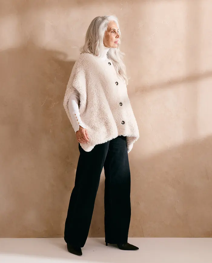 Mulher com casaco poncho creme e calças pretas em fundo bege