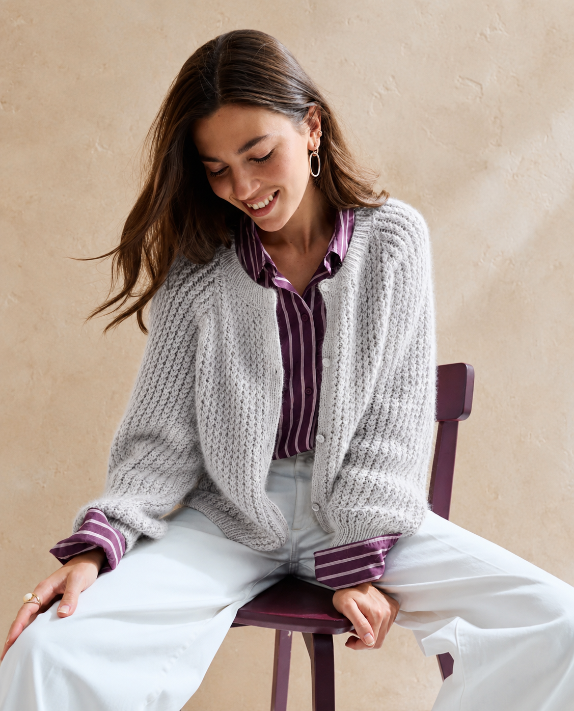 Mulher com cardigan cinzento, camisa roxa às riscas brancas e calças brancas sentada numa cadeira roxa