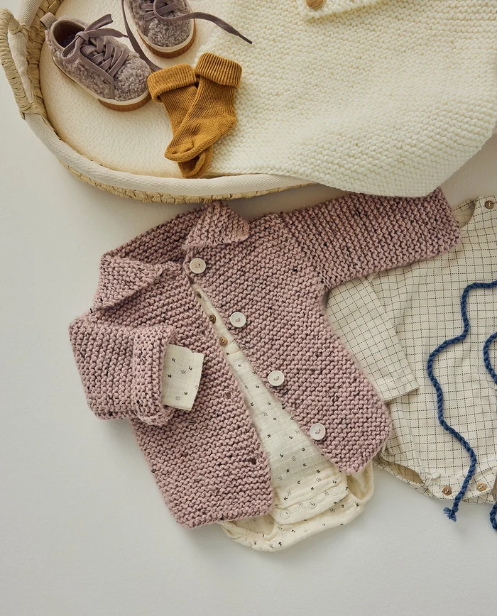 Conjunto de roupa de bebé com casaco rosa tricotado, body e camisa padrão quadriculado, meias castanhas e sapatos castanhos felpudos num berço com manta creme.