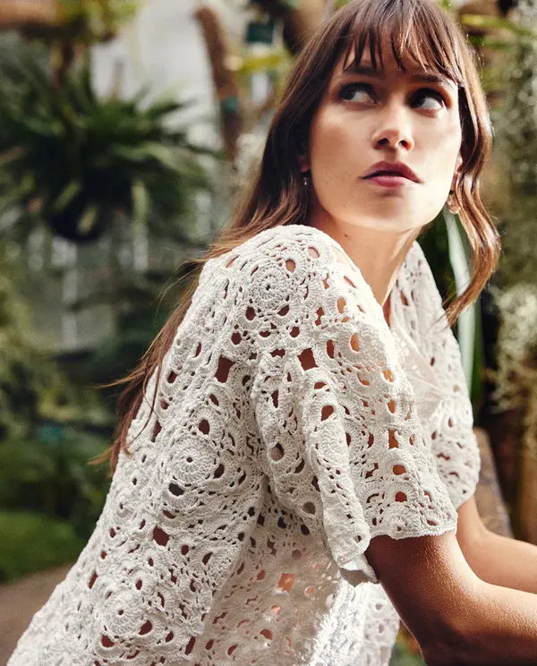 Mulher de perfil a usar blusa branca de croché com padrão floral, em ambiente exterior com plantas ao fundo.