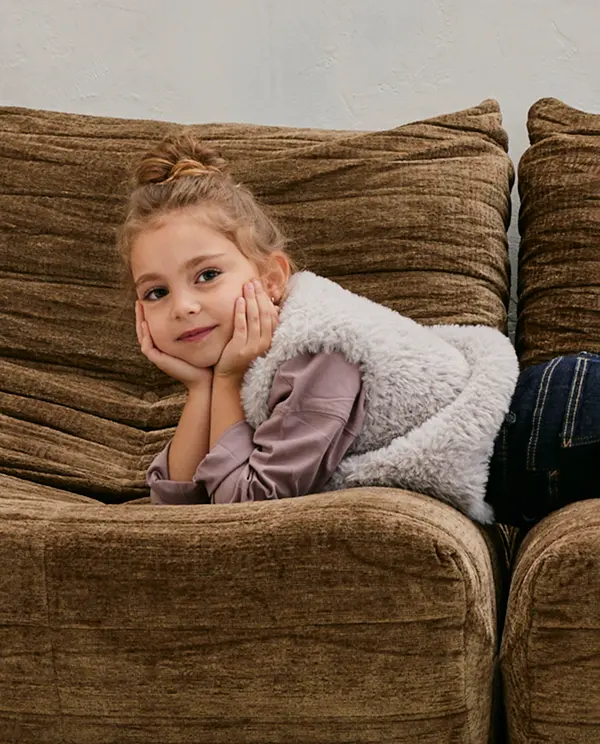 Criança deitada em sofá castanho usando colete branco de pelo sintético e camisa lilás