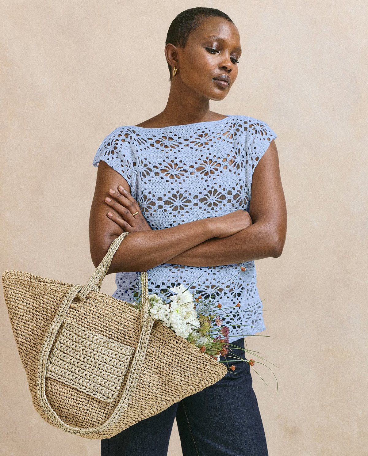 Mulher com camiseta azul clara de crochê, calças azuis, e bolsa de palha com flores