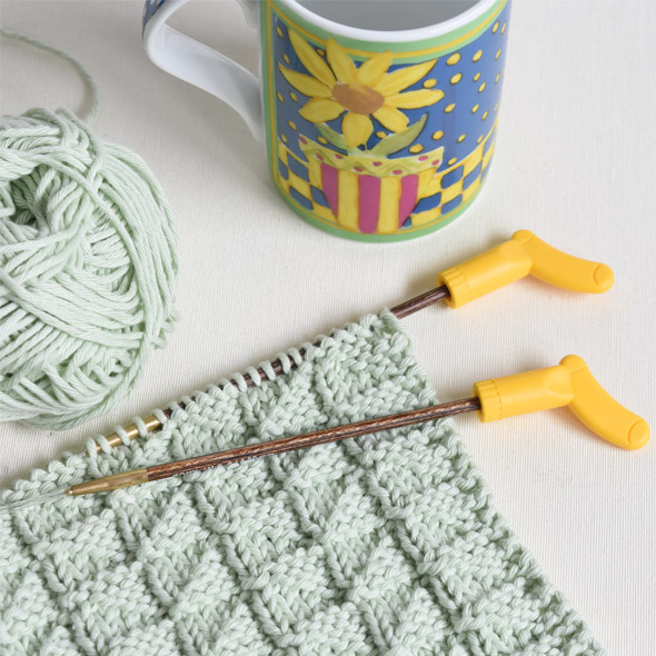 Peça de tricô verde claro, agulhas metálicas amarelas, novelo de lã e caneca colorida